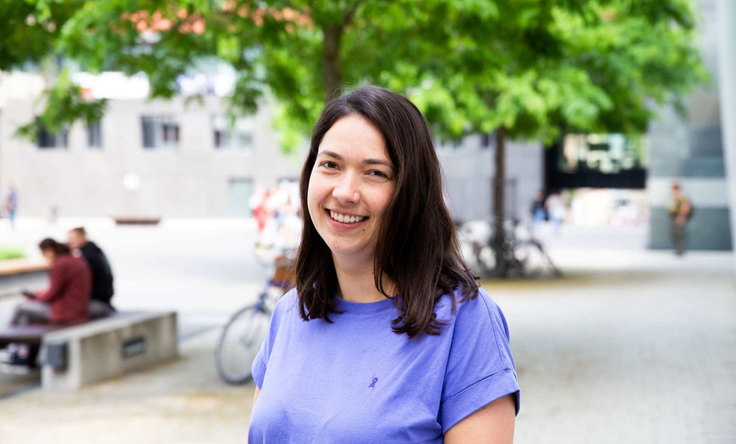 Eine Frau mit schulterlangen, dunkelbraunen Haaren und einem blauen T-Shirt steht auf einem großen Hof mit Bäumen und Sitzbänken und lächelt in die Kamera.