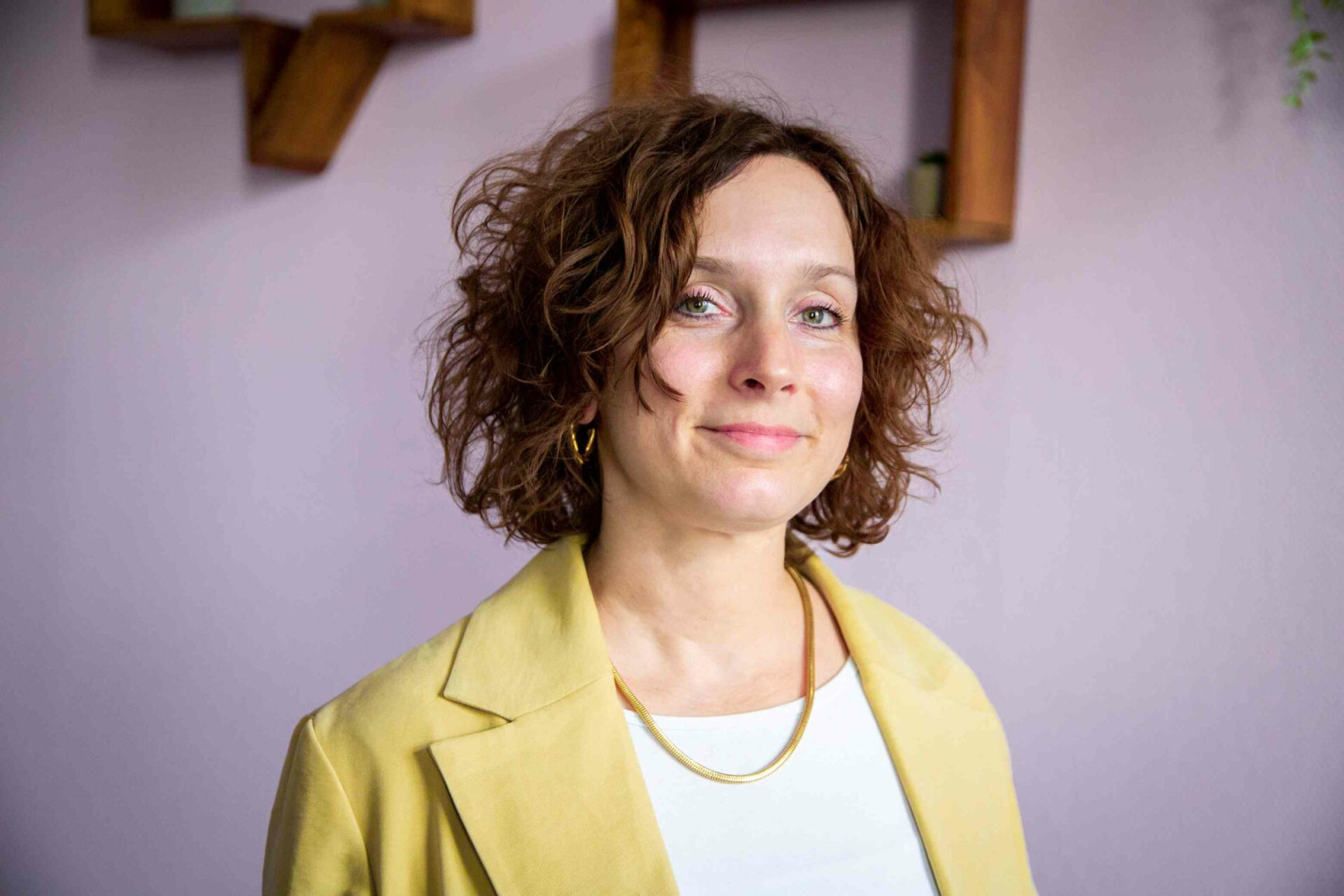 Eine Frau mit kinnlangen, braun-gelockten Haaren, einem gelben Blazer und einer goldenen Kette blickt lächelnd in die Kamera.