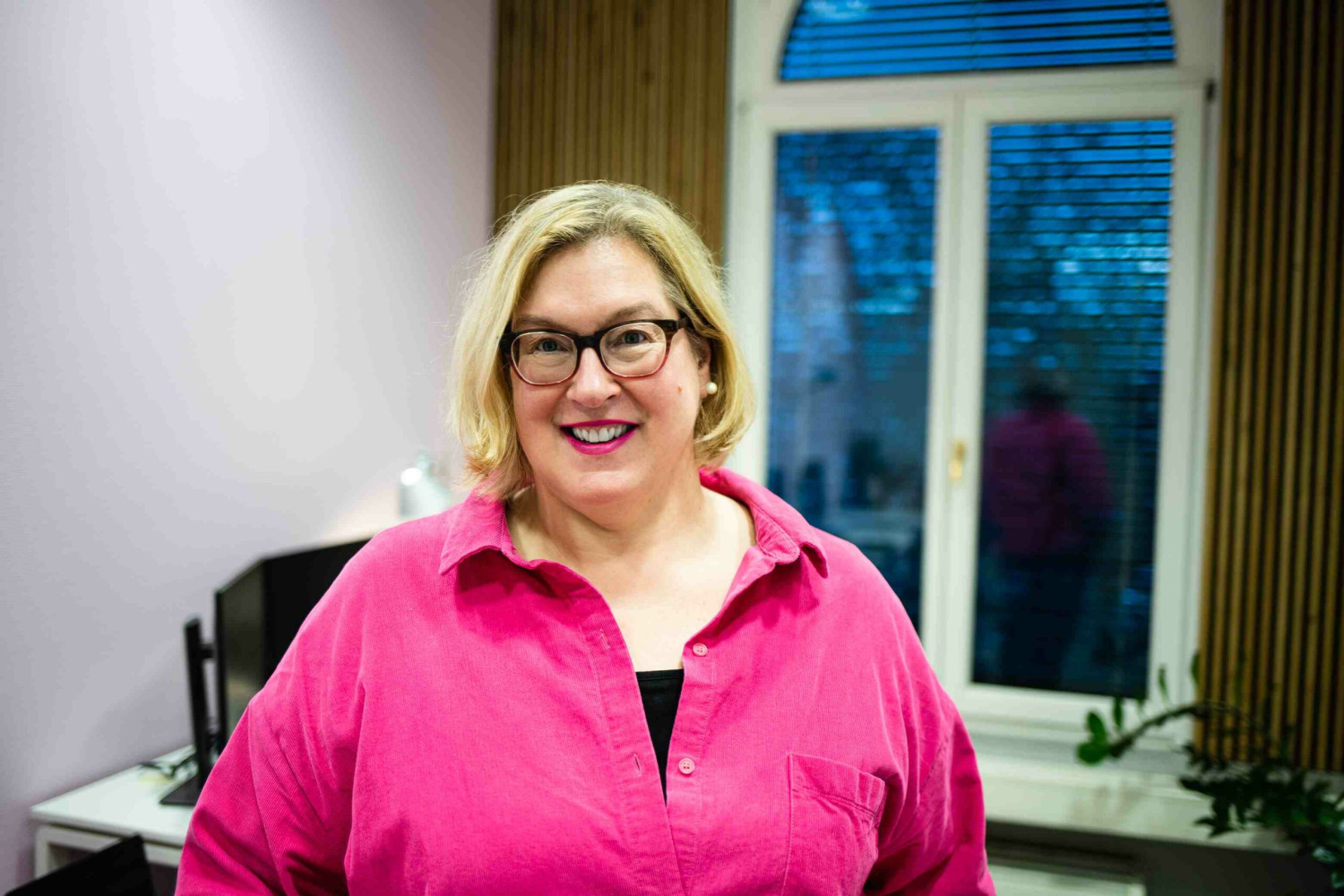 Eine blonde Frau mit dunkler Brille und einer rosafarbenen Bluse steht in einem Büro und lächelt in die Kamera.
