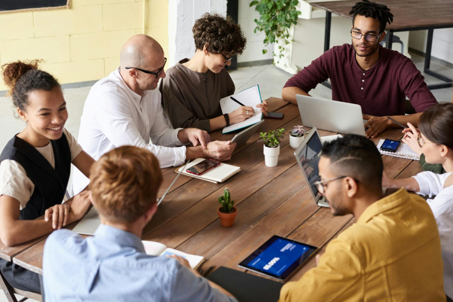 Sechs Personen sitzen an einem Tisch in einem modernen Büro mit Laptops, Tablets und Notizbüchern