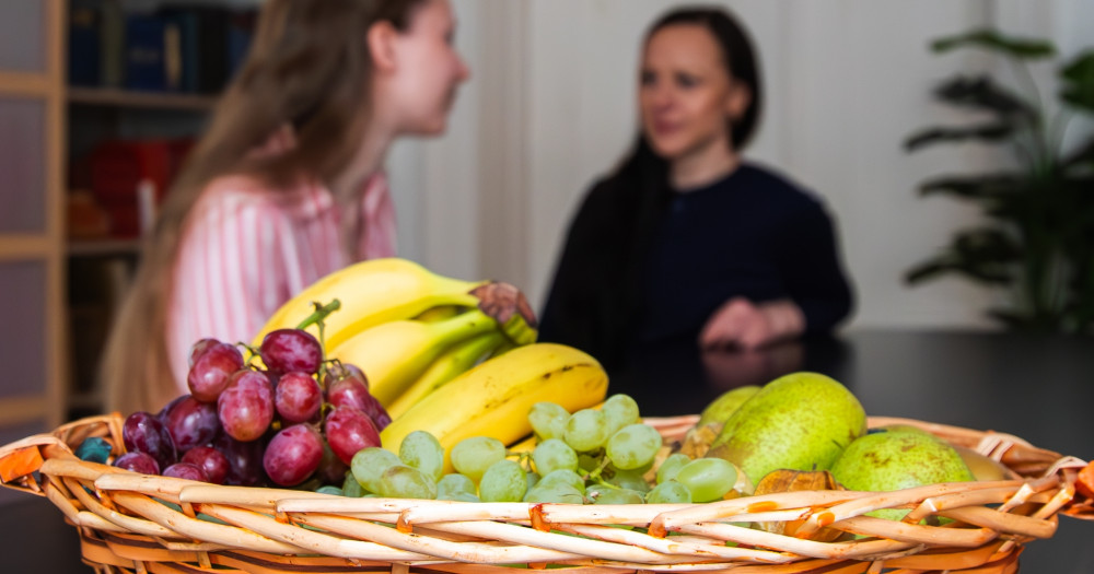 Zwei Frauen sitzen an einem Tisch, im Vordergrund steht ein Obstkorb.