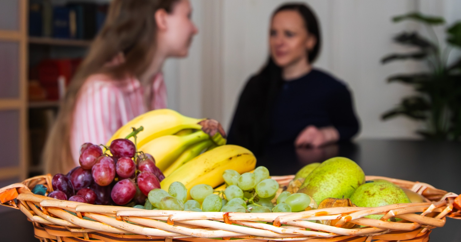 Zwei Frauen sitzen an einem Tisch, im Vordergrund steht ein Obstkorb.