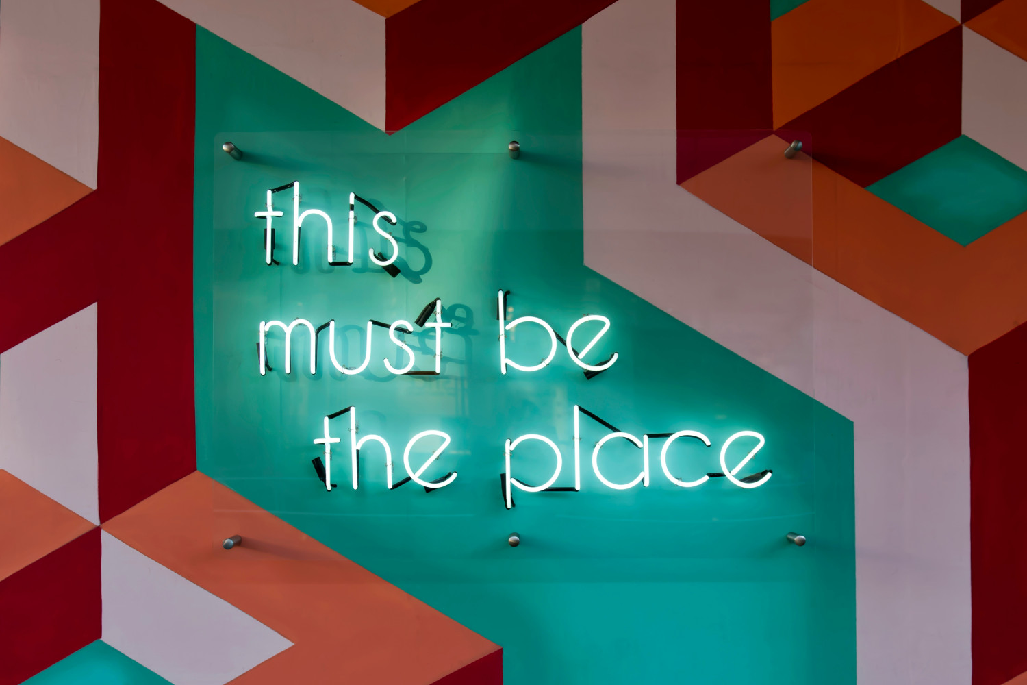 Neon-Schriftzug ‚this must be the place‘ auf einer Glasplatte vor einer geometrisch gemusterten Wand in Türkis, Orange und Weiß