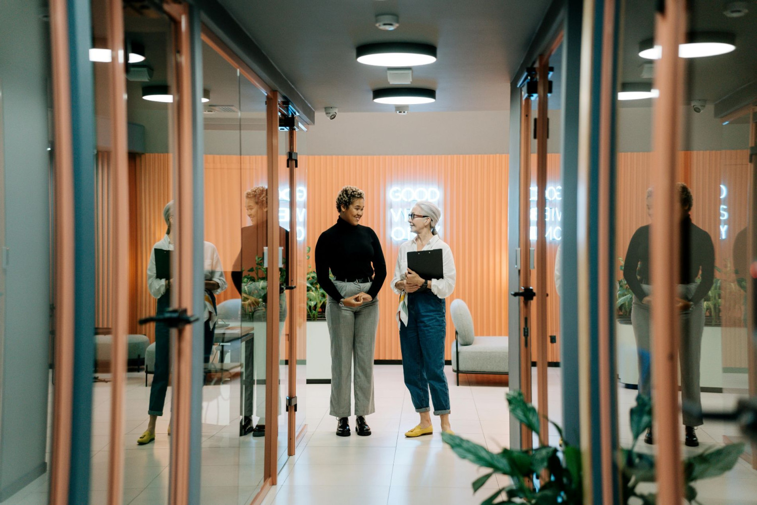 Zwei Frauen unterhalten sich lächelnd in einem modernen Büroflur mit Glaswänden.