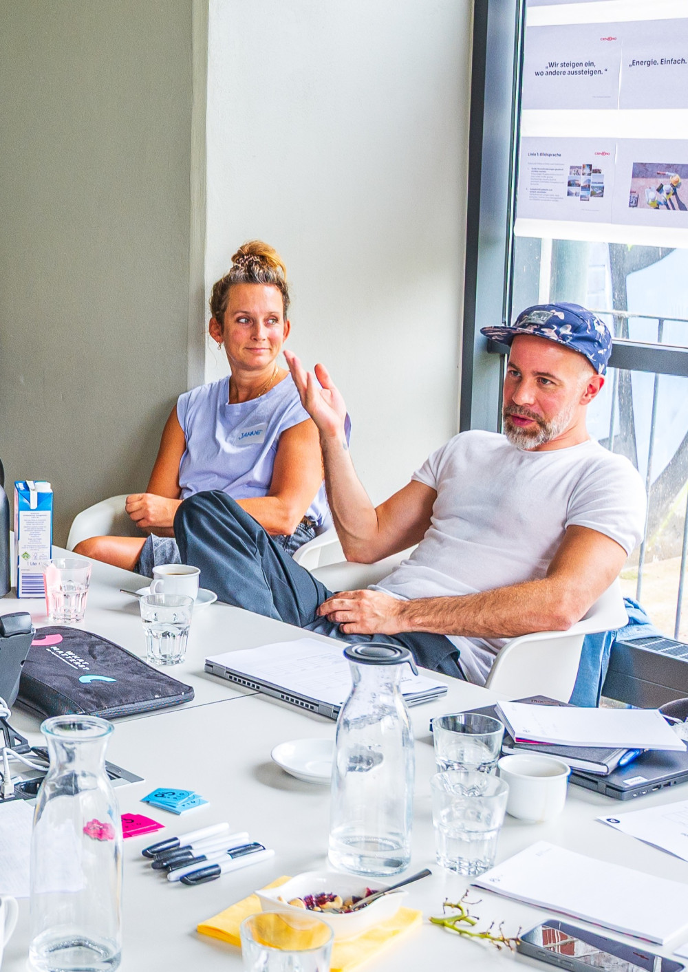 Eine Frau und ein Mann sitzen in einem Workshop. Vor ihnen auf dem Tisch befinden sich Laptops, Getränke und Kommunikationsmaterialien.