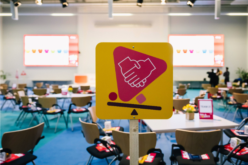 Ein leerer Saal mit Tischen und Stühlen. Im Vordergrund befindet sich ein gelbes Schild mit einem rosafarbenen Symbol zweier Hände, die sich wie zur Begrüßung schütteln.