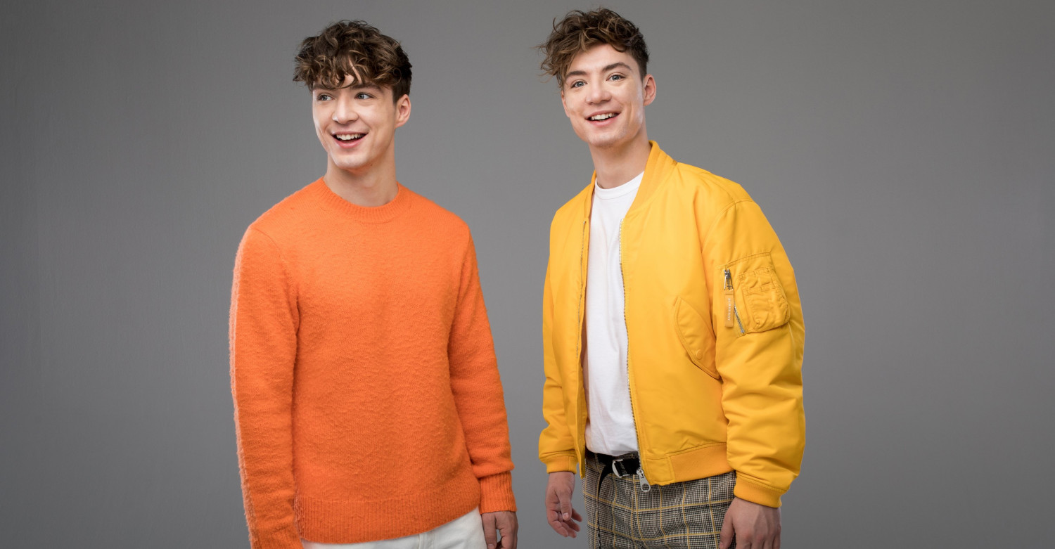 Zwei junge Männer mit lockigen, kurzen Haaren und einem orangefarbenen Pulli beziehungsweise einer gelber Jacke