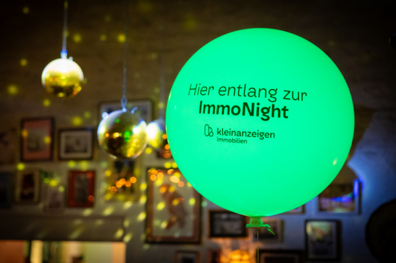 Grüner Luftballon mit der Aufschrift "Hier entlang zur ImmoNight" von Kleinanzeigen Immobilien. Im Hintegrund befinden sich Diskokugeln
