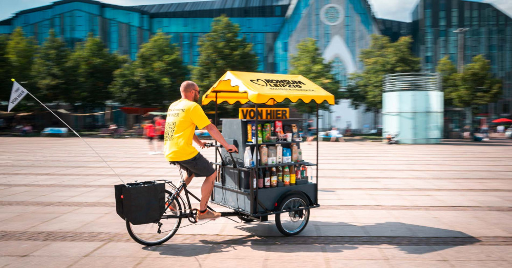 Mann fährt mit gelbem Konsum-T-Shirt und mobilem Supermarkt auf dem Fahrrad über den Augustusplatz in Leipzig