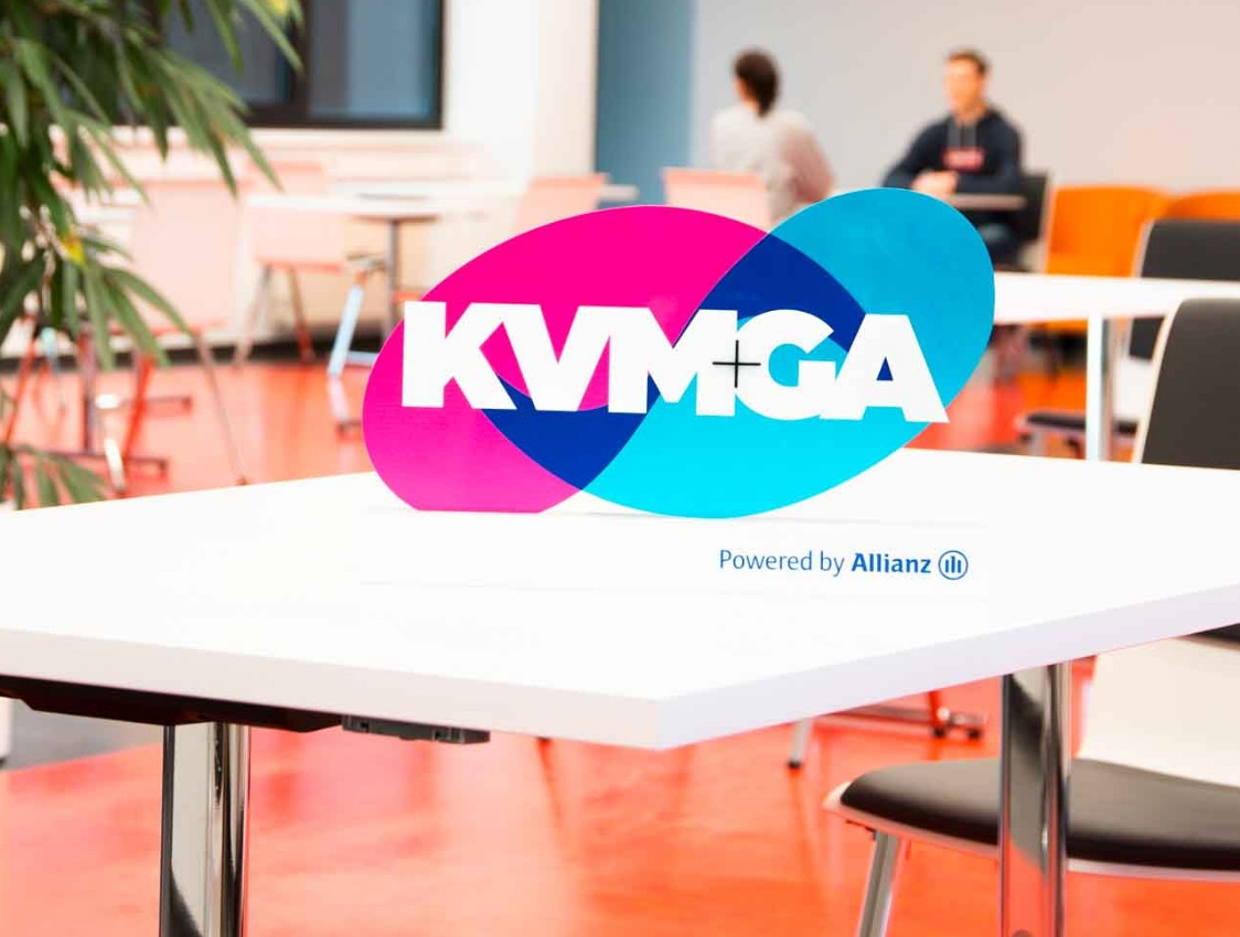 Logo KVM +GA auf Hintergrund mit zwei Personen und einem Schreibtisch.
