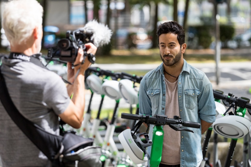 A cameraman films a man with black hair and a denim jacket in front of a row of Lime E-scooters