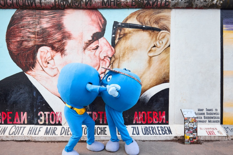 Zwei Pointies, die runden, blauen Maskottchen von PayBack, küssen sich vor dem Graffiti des berühmten sozialistischen Bruderkuss' von Dmitri Wladimirowitsch Wrubel in Berlin.