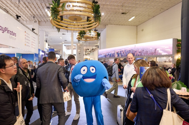 Ein Pointie, das runde, blaue Maskottchen von PayBack läuft durch die Gänge der Messe, beobachtet von zahlreichen anderen Besucher*innen.