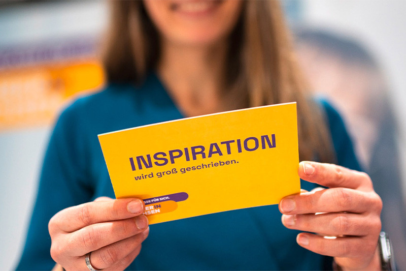 Frau, die eine Postkarte mit der Aufschrift "INSPIRATION wird groß geschrieben" in den Händen hält.