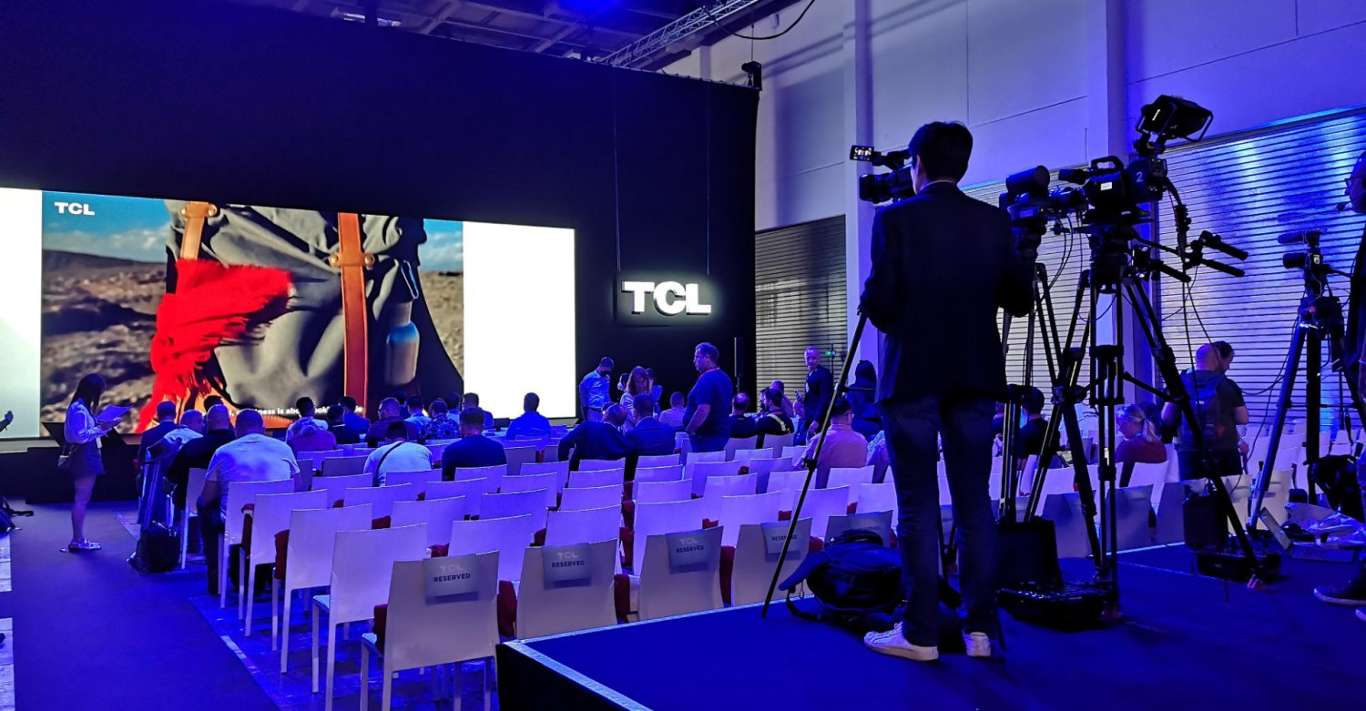 Ein Kameramann filmt eine Produktvorstellung von TCL auf der Messe. Das Publikum sitzt auf Stühlen vor einer großen Leinwand.