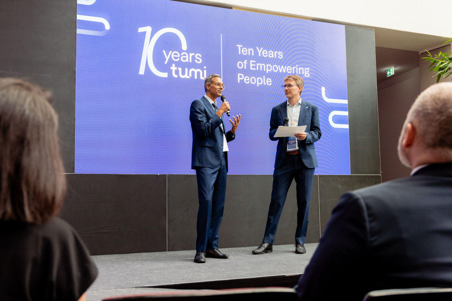 Two men in suits stand on a stage in front of a large screen displaying the text '10 years tumi Ten Years of Empowering People' speaking to an audience.