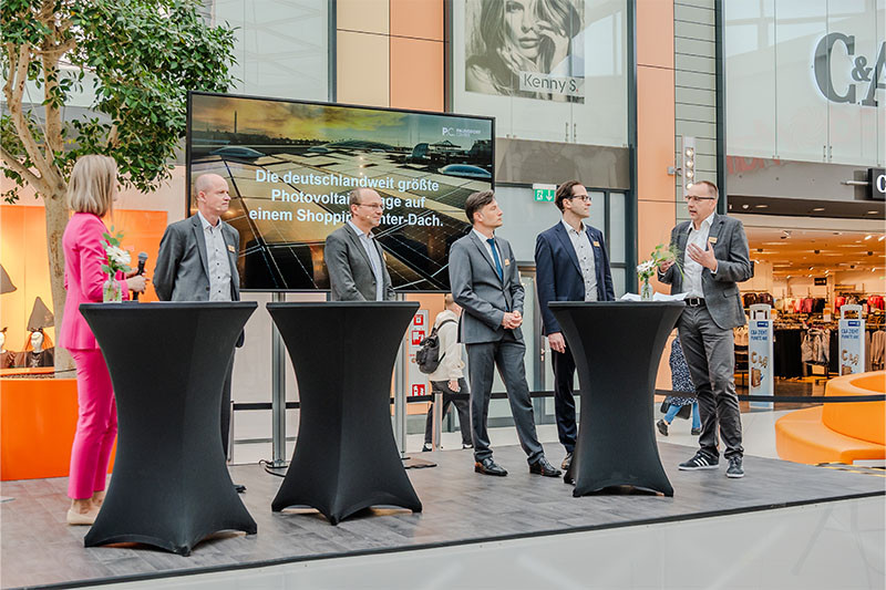 Several festively dressed people are standing at bar tables in a shopping center. Behind them is a large monitor that reads “Germany's largest photovoltaic system on a shopping center roof”.