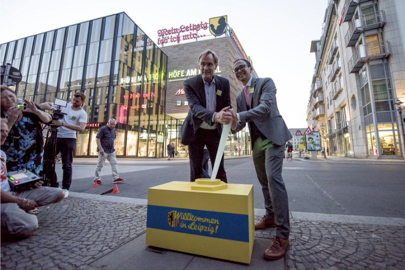 Two men pull a large gear lever in front of the “Höfe am Brühl” shopping center in Leipzig. The scene is photographed and filmed by several people.