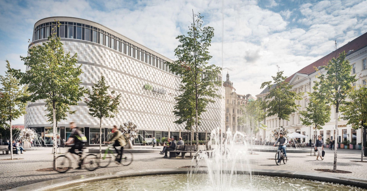 Blick auf das Einkaufszentrum "Höfe am Brühl" in Leipzig, mit einem Springbrunnen und Bäumen im Vordergrund