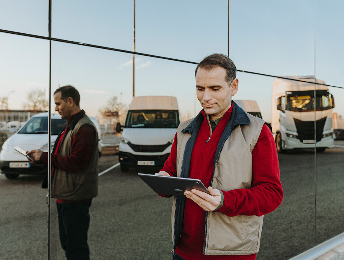Ein Mann steht vor einer Fassade, in dem sich verschiedene Fahrzeuge vom PKW bis zum LKW spiegeln. In der Hand hält er ein Tablet.