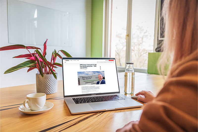 Eine Frau sitzt an einem Tisch mit einem Laptop und liest eine Meldung von Webfleet. Neben dem Laptop stehen eine Kaffeetasse, eine Wasserflasche und eine Grünpflanze.