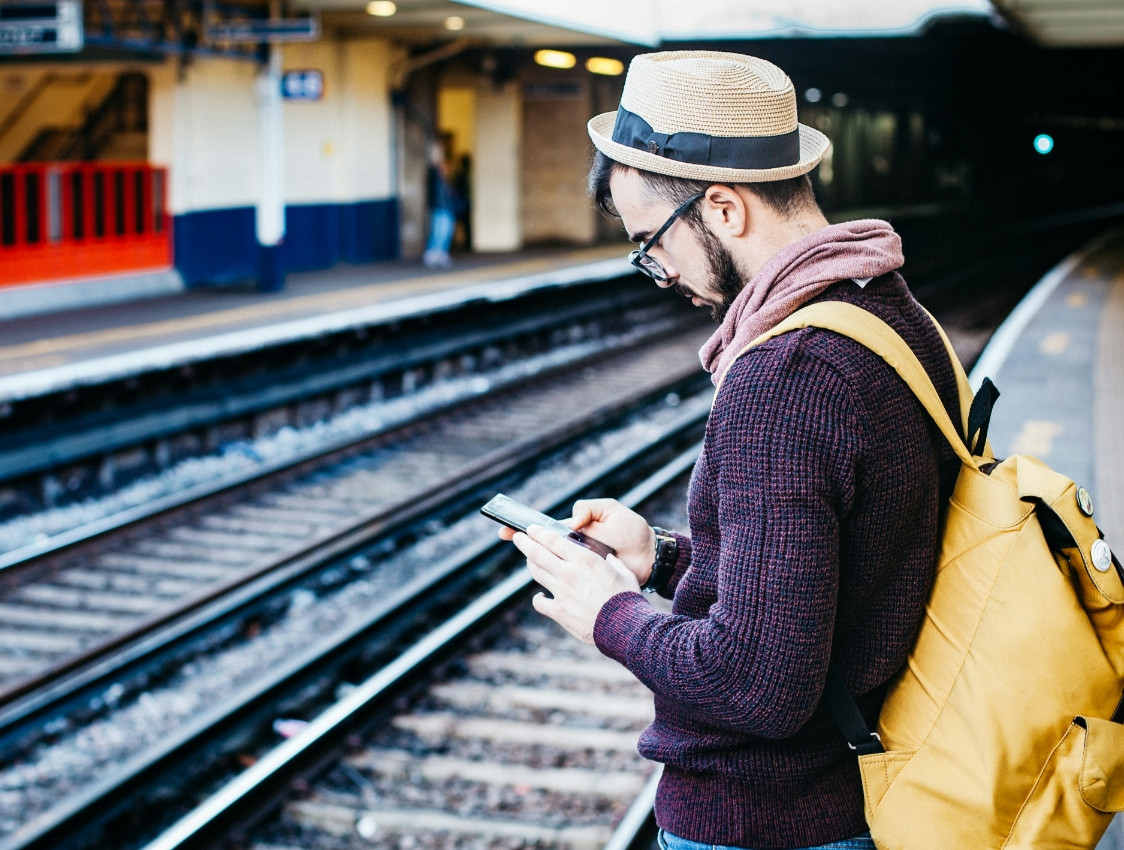 Ein Mann mit Hut und Rucksack steht an einem Bahngleis und blickt auf sein Smartphone