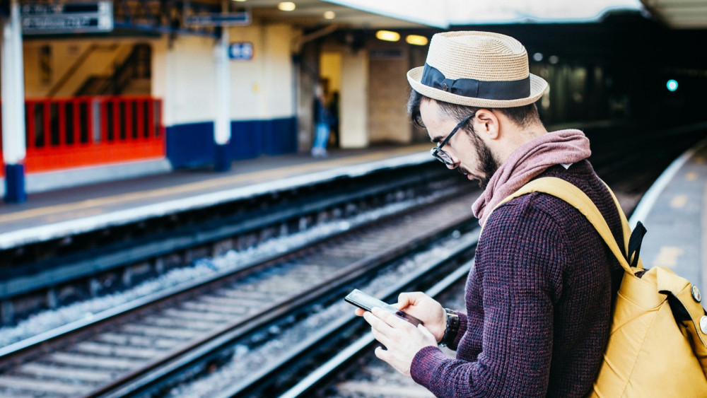 Ein Mann mit Hut und Rucksack steht an einem Bahngleis und blickt auf sein Smartphone