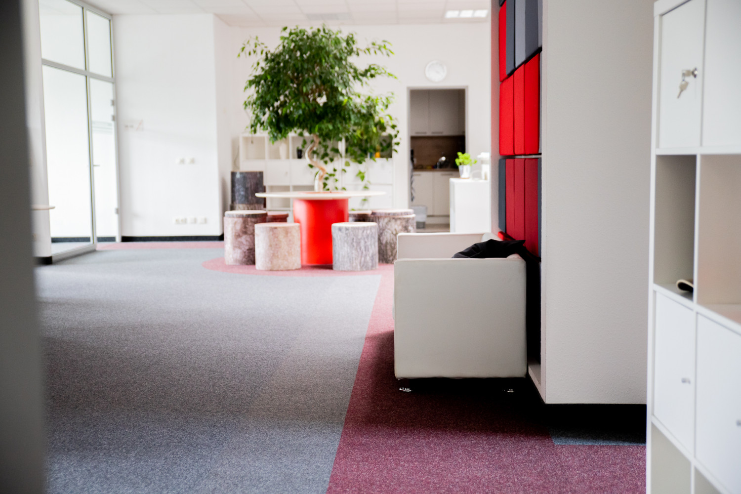 Ein helles, offenes Büro mit einem runden, roten Tisch, in dessen Mitte ein Baum steht. Die Stühle sind Holzstämme. Auf der rechten Seite befinden sich Regale mit roten und grauen Fächern und ein weißer Sessel. Den Boden bedeckt ein grau-roter Teppich.