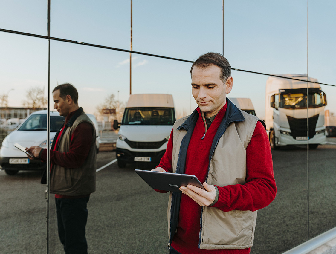 Ein Fuhrparkmanager mit einem Tablet mit der Software von Webfleet in der Hand vor einer betrieblichen LKW-Flotte.